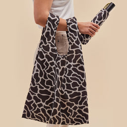 Person holding a tote bag with black and white ripples pattern, holding an umbrella against a beige background