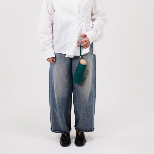Person wearing a white shirt and blue jeans holding a mini green umbrella on a plain background