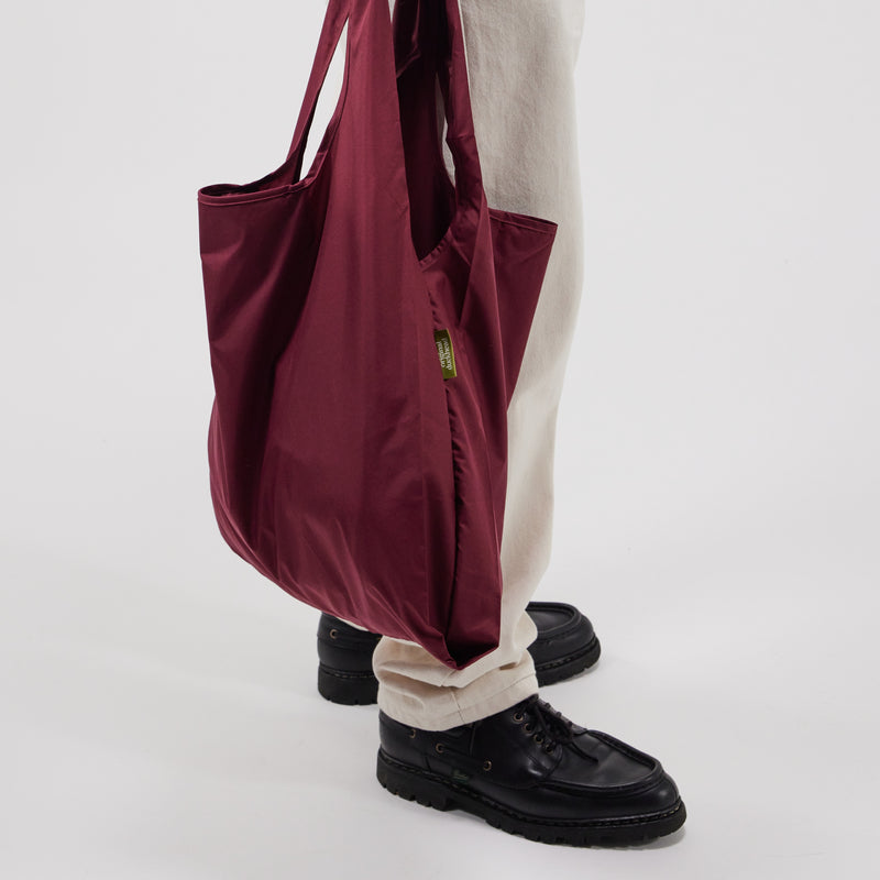 Cherry red tote bag held by a person wearing black shoes and beige pants on a white background